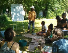 Photographie de personnes assises en cercle dans l'herbe devant une personne debout à côté d'un tableau